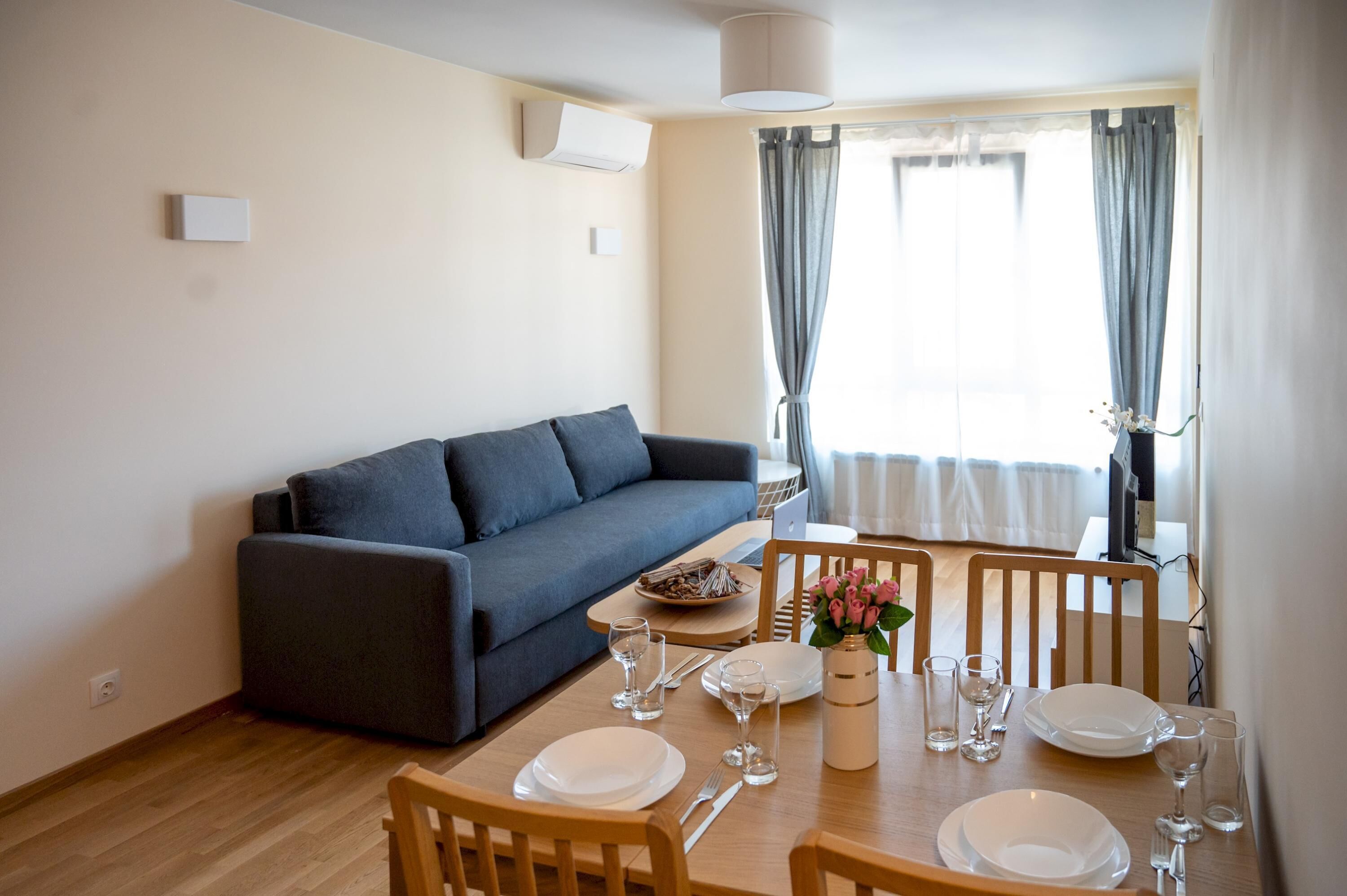 Un comedor con mesa de madera dispuesta para cuatro personas, sofá gris y ventana con cortinas.