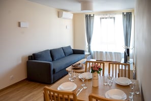 A dining area with a wooden table set for four, a grey sofa, and a window with curtains. - Spacious 1bdr Apartment in City Centre With Balcony (Sofia)