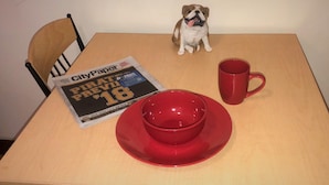 A newspaper titled "City Paper" with a date of "2018" and a headline about a "Field Fair". A red mug and a red bowl are on the table.