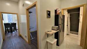 Camera d'albergo con angolo cottura, un piccolo tavolo con un tostapane, un frigorifero e un cestino. Si vede una porta che conduce a un bagno con doccia e un asciugamano appeso al muro.