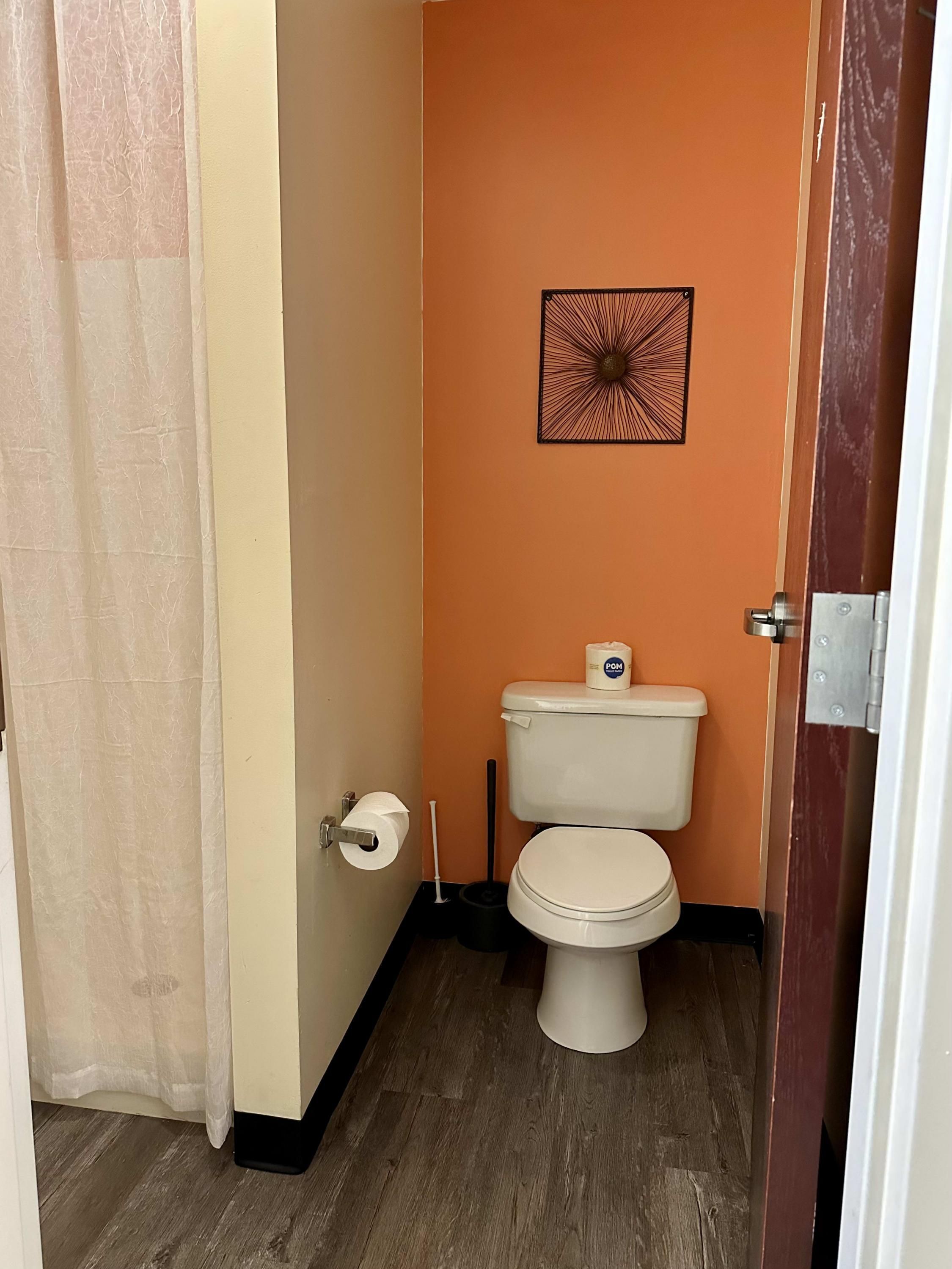 A small bathroom with an orange wall, a toilet, a roll of toilet paper, and a black cleaning brush.