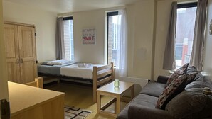Una habitación pequeña y bien iluminada con una cama, un sofá, una mesa de madera y un armario. Hay un letrero en la pared que dice “BE HAPPY LAUGH OUT LOUD ACT SILLY SMILE DAYDREAM”.