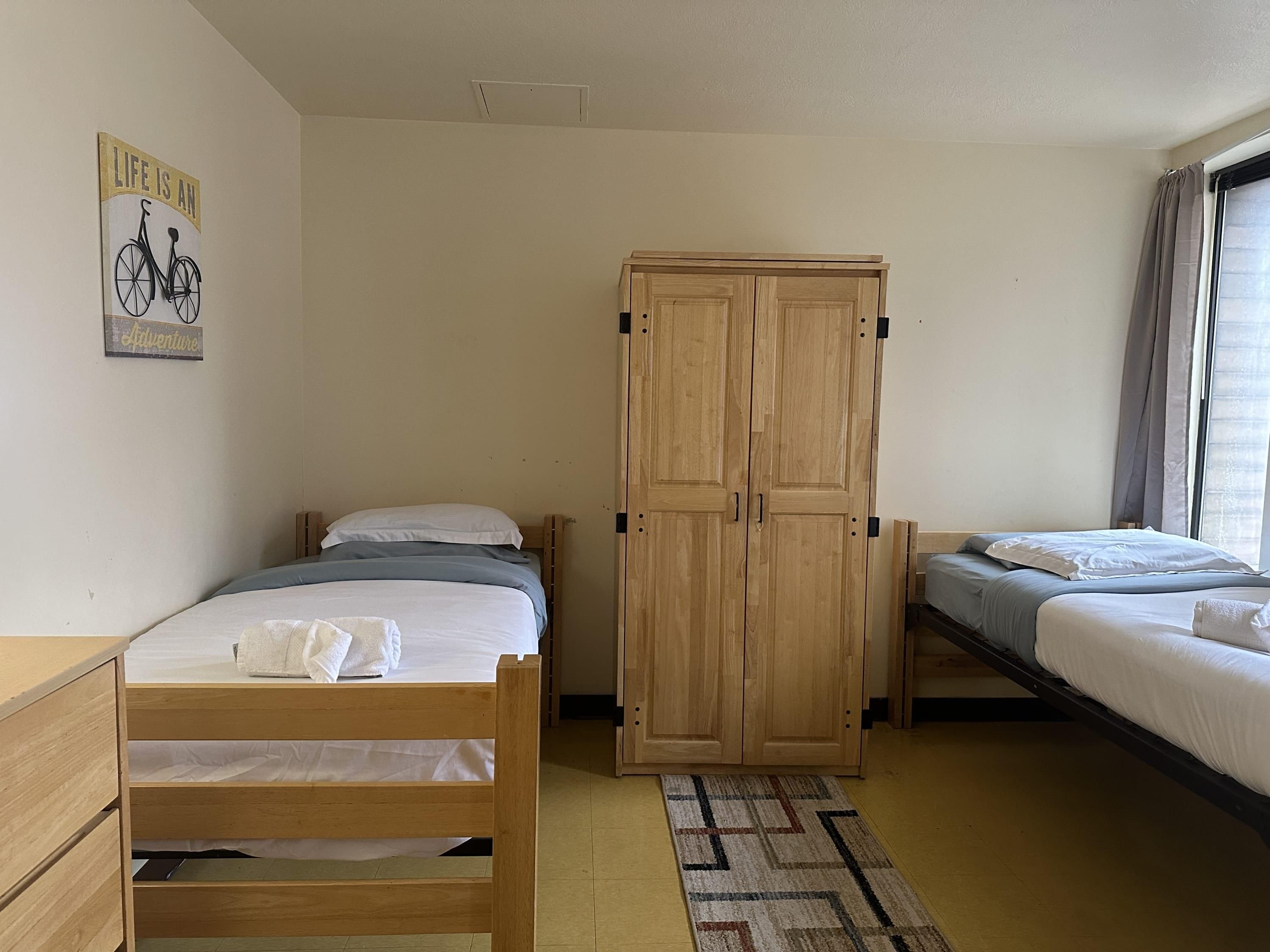 Ein Zimmer mit zwei Betten, einem Holzschrank, einem kleinen Schreibtisch und einem Poster an der Wand.