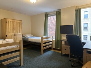 Quarto de dormitório com duas beliches, um guarda-roupa de madeira, uma escrivaninha com cadeira, uma televisão e uma janela com cortinas.