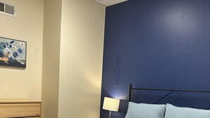 Una cama bien hecha con estructura metálica, almohada azul claro y colcha blanca en una habitación con pared de acento azul oscuro y cabecera de madera.