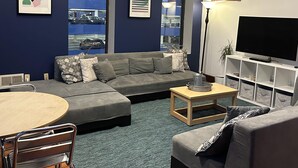 Una sala moderna con un sofá seccional gris, una mesa redonda de madera, un televisor sobre un estante blanco y ventanas grandes con vistas a la ciudad.