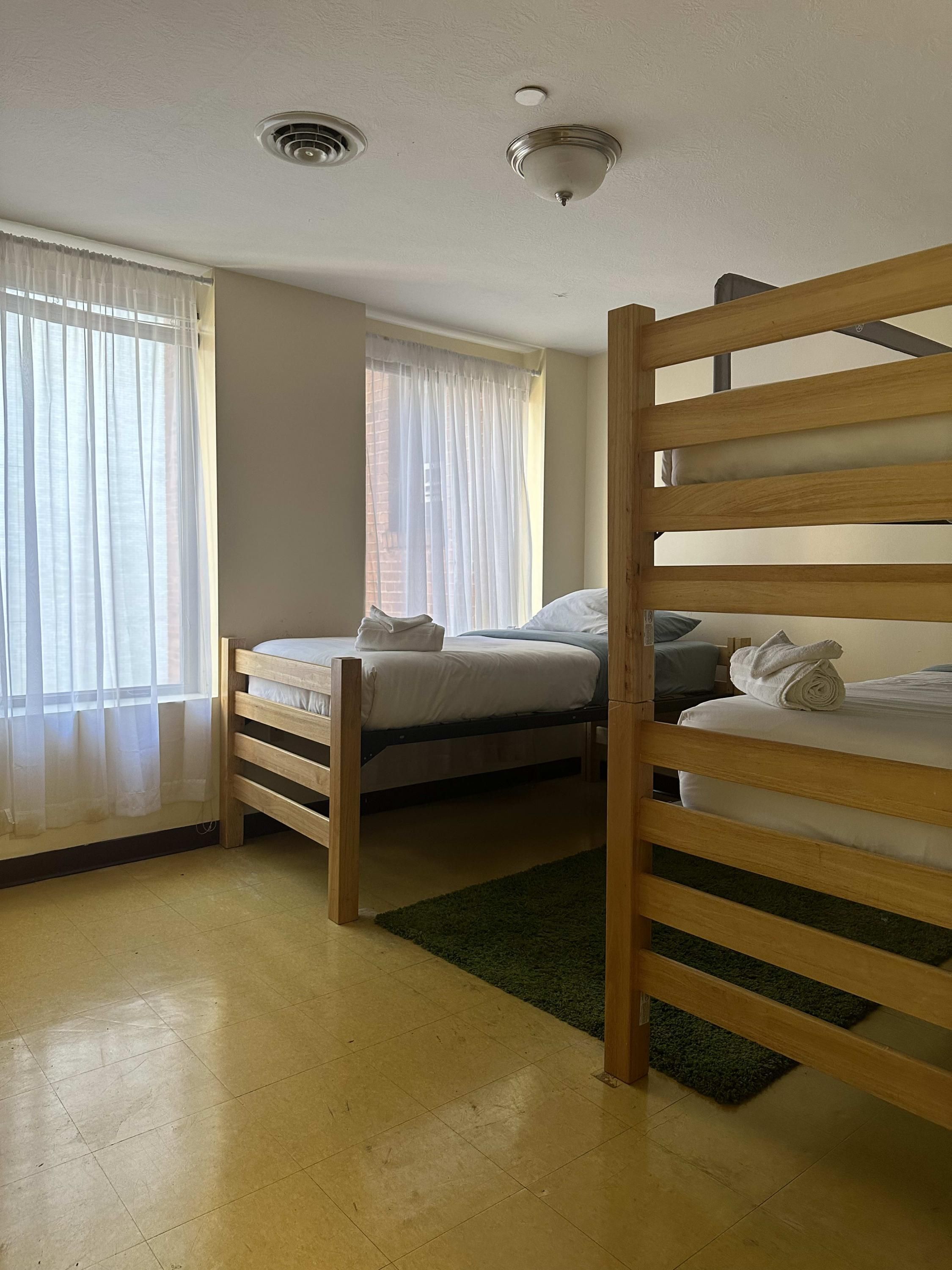 A room with two bunk beds, a window with sheer curtains, a ceiling vent, and a green rug.