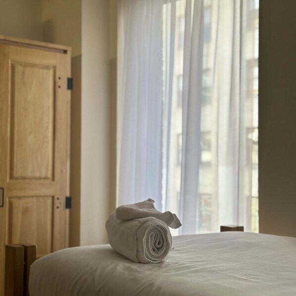 Una cama bien hecha con una toalla enrollada encima, un armario de madera y una ventana con cortinas traslúcidas.