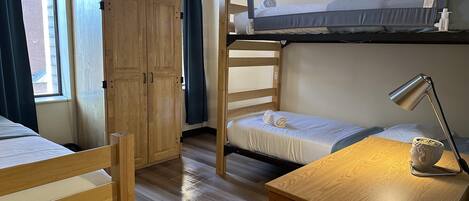 Un dormitorio pequeño con literas, un armario de madera, un escritorio con lámpara y una ventana con cortinas.