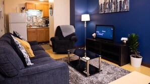 A living room with a dark blue wall, a cityscape painting, a television, a sofa, a chair, a small table, and a kitchen in the background. - 2 Bedroom Downtown Apt Clean and Safe (Pittsburgh)