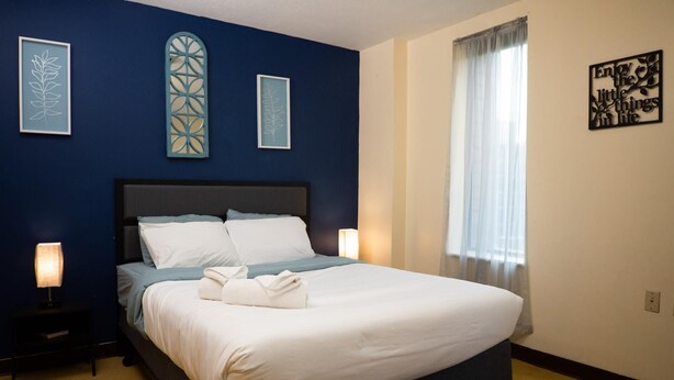 A neatly made bed with white linens and a blue accent, a bedside lamp, and a framed artwork on the wall. - 2 Bedroom Downtown Apt Clean and Safe (Pittsburgh)