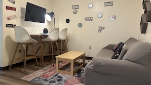 Ein kleiner, gemütlicher Raum mit einem Holztisch, zwei weißen Stühlen, einem grauen Sofa und einem an der Wand befestigten Flachbildfernseher.