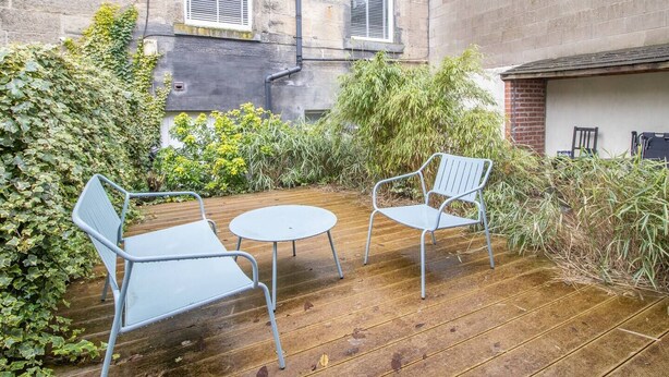 A wooden deck with two metal chairs and a small table, surrounded by greenery and a stone building with windows. - Stylish West End Detached Mews - Pass the Keys (Glasgow)