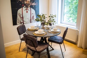 Una mesa de comedor dispuesta con platos, tazones y vasos, rodeada de sillas, con una ventana y un cuadro al fondo.