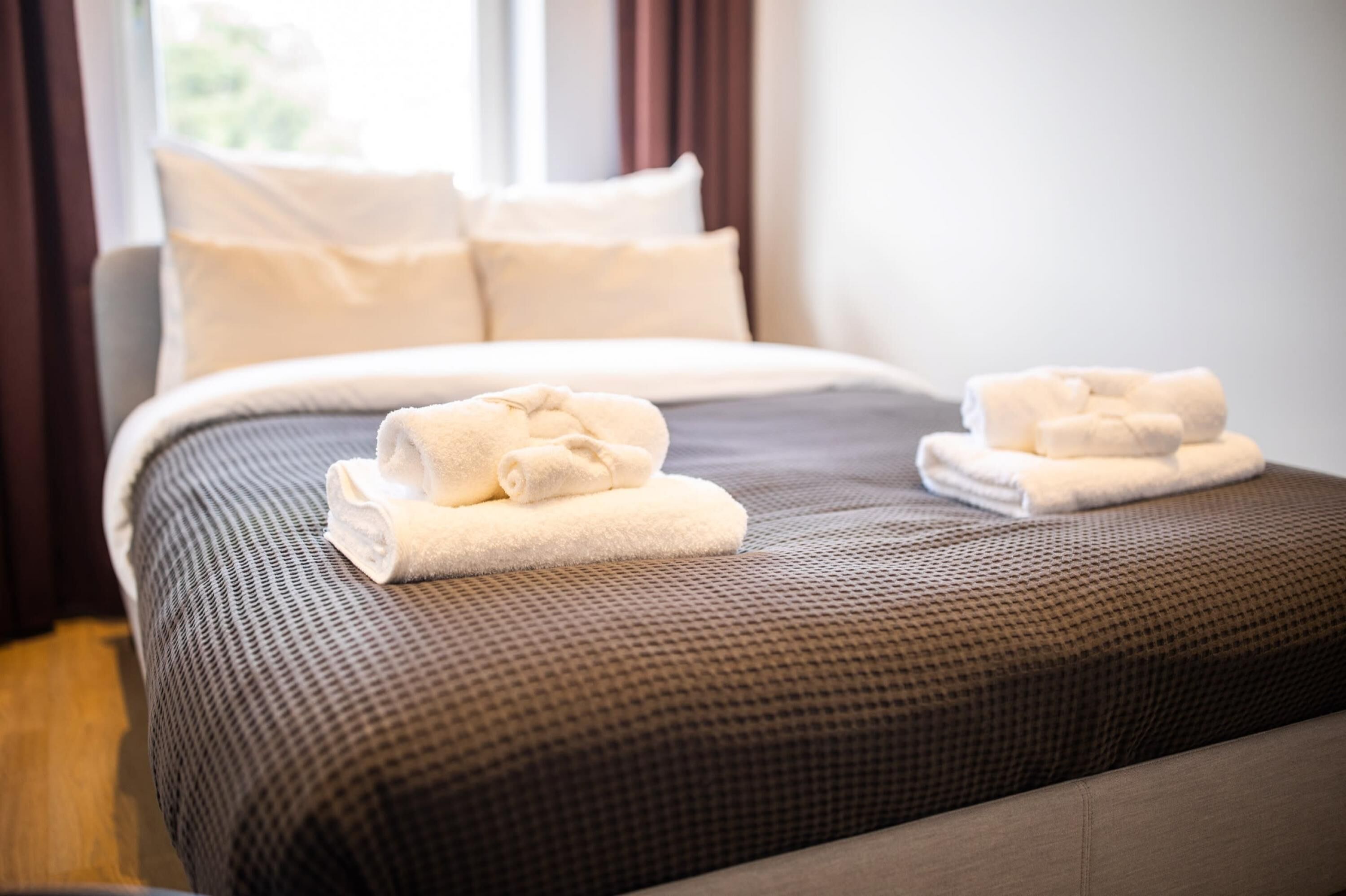 A neatly made bed with a brown textured bedspread and two rolled white towels placed on the footboard.