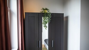 Ein dunkler Holzschrank mit Pflanze obenauf, ein Bett mit weißem Kopfteil und ein Fenster mit Vorhängen.