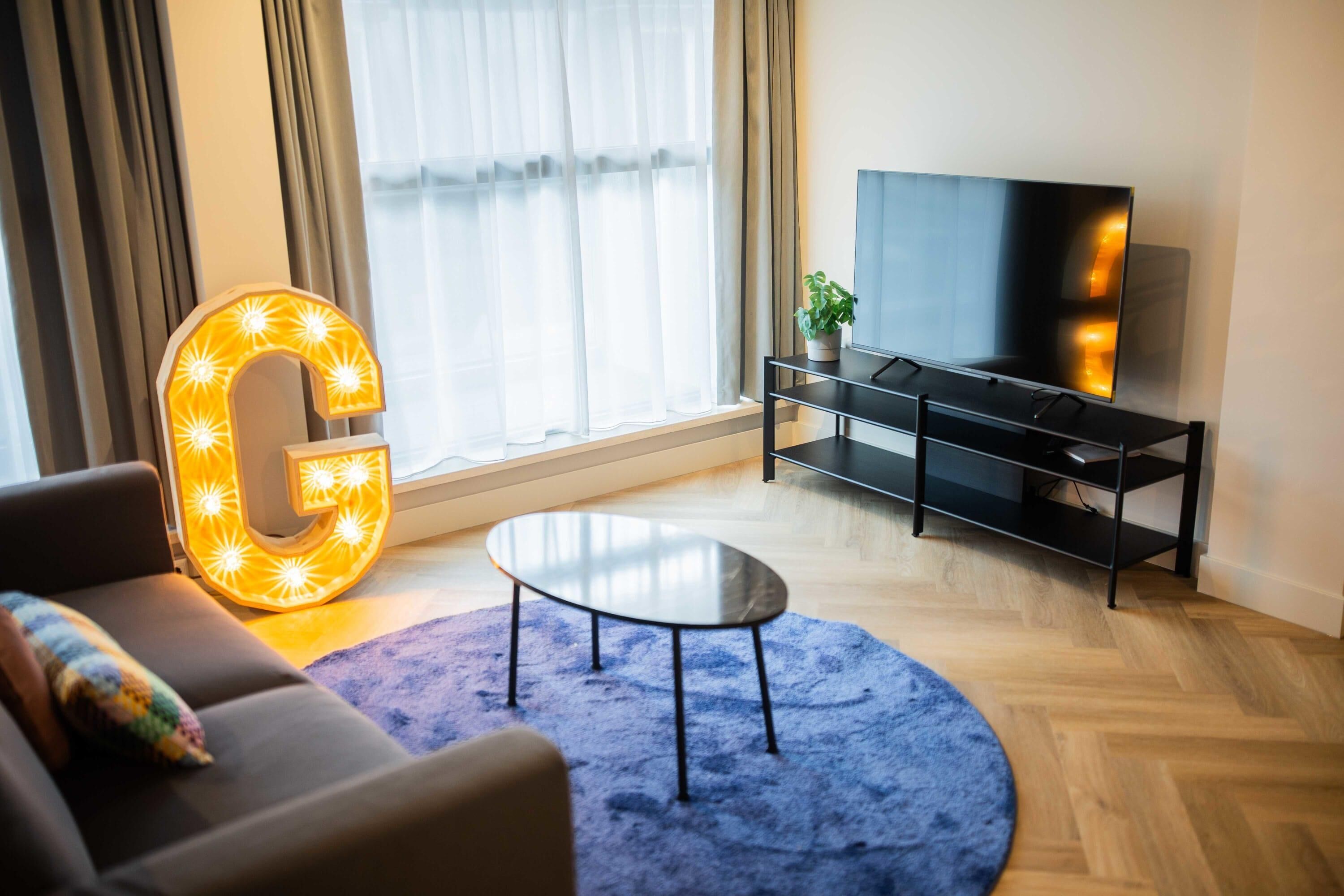 A modern living room with a large illuminated 'G' sign, a flat-screen TV, a sofa, a coffee table, and a potted plant.