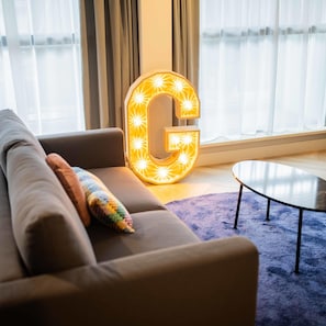 A modern living room with a large illuminated 'G' sign, a sofa with a colorful pillow, and a round coffee table. - Peaceful Serviced Studio's -44m2- (Hilversum)