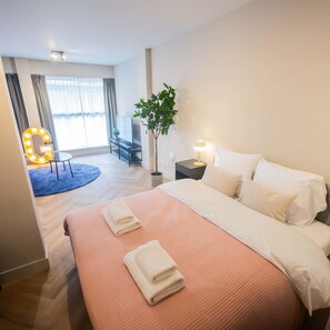 Ett hotellrum med en stor säng, en TV på ett stativ, ett litet bord med en växt, en lampa och en spegel med inbyggd belysning.
