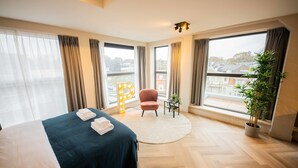 Een hotelkamer met een groot bed, een rond vloerkleed, een roze stoel, een potplant en uitzicht op de stad door grote ramen.