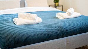 A neatly made bed with blue bedding and white pillows, a small bedside table with a plant, and a window with curtains. - Serviced 1BR Apartments in Utrecht (De Bilt)