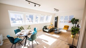 A spacious living area with a dining table, chairs, a sofa, and a coffee table. There are large windows providing natural light and a view of the outside. - Serviced 1BR Apartments in Utrecht (De Bilt)