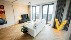 Una sala de estar moderna con un sofá blanco, un televisor sobre un soporte negro, una mesita auxiliar con una planta y un gran letrero iluminado con la letra 'V'.