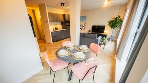 A modern apartment with a dining area featuring a round table set for four, a kitchen in the background, a living area with a sofa and TV, and a large window providing natural light. - Joyful Serviced Studio Apartment -ks71-j (Hilversum)