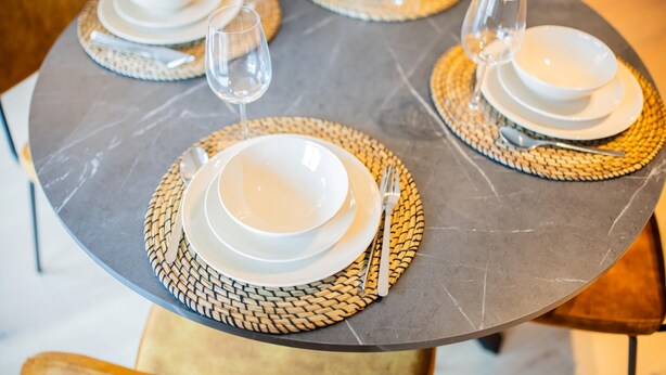 A table set with white plates, bowls, and cutlery, accompanied by woven placemats and wine glasses. - Alphabet 3 Bedroom Serviced Apartment -pk- (Rotterdam)