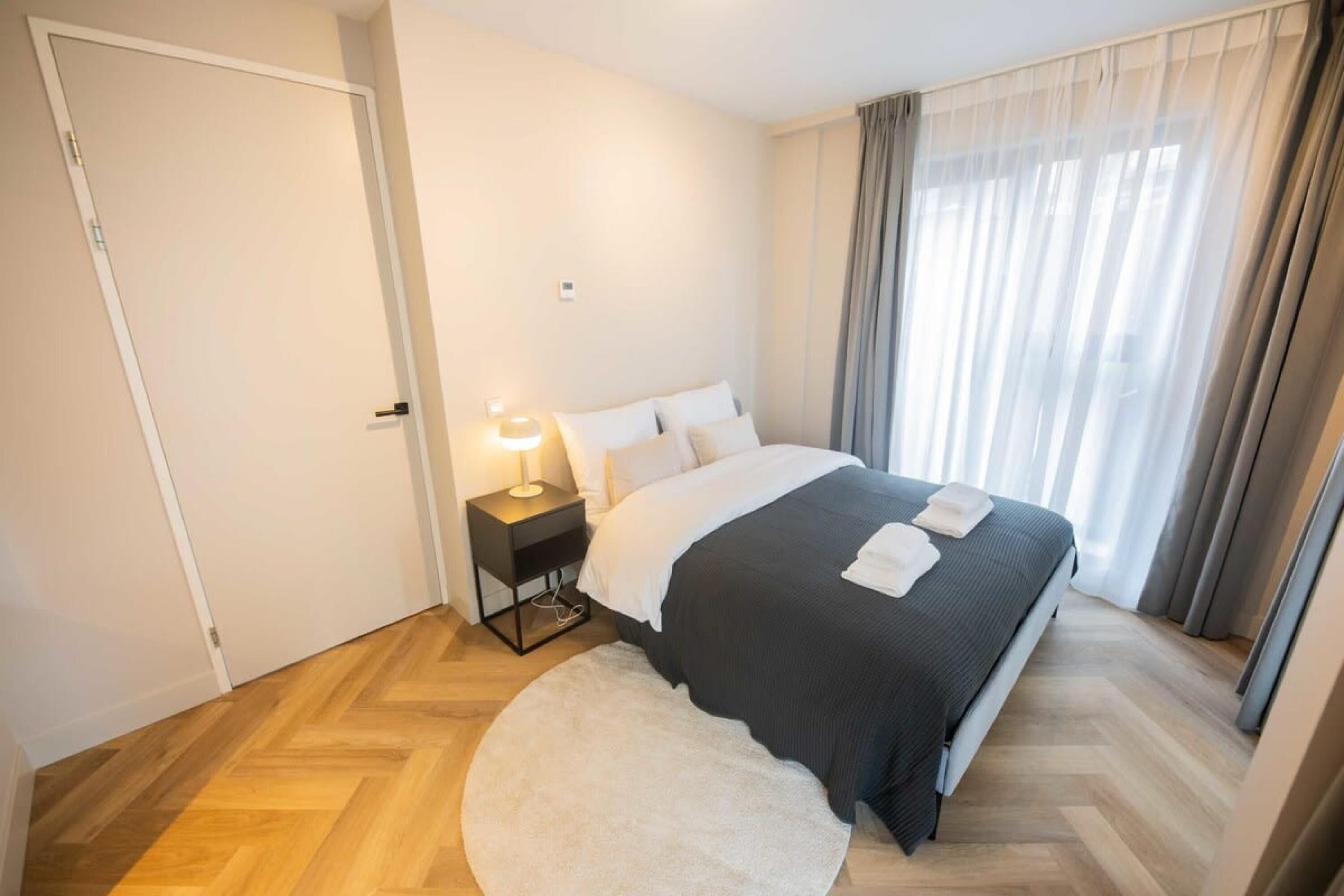 Une chambre à coucher avec un lit, une table de chevet avec une lampe, un pouf rond blanc et une fenêtre avec des rideaux.