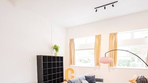 A modern living room with a grey sofa, a wooden dining table, a black shelving unit, and a large window with curtains. - Serviced 3BR Apartment in Utrecht (De Bilt)