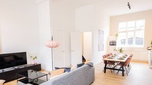A modern living room with a grey sofa, a flat-screen TV, a pink floor lamp, and a dining area with a wooden table and chairs. - Serviced 3BR Apartment in Utrecht (De Bilt)
