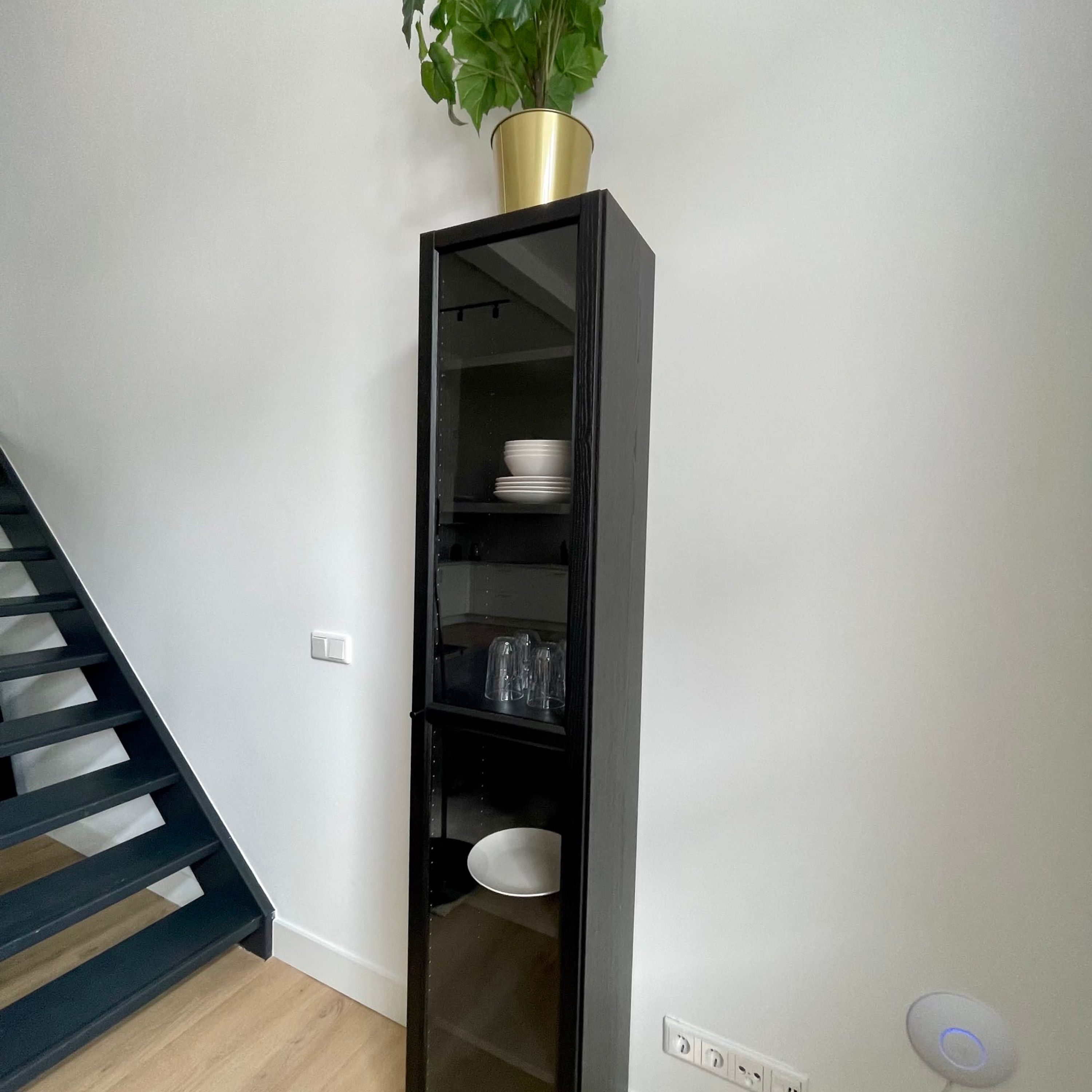 Une étagère noire sur laquelle sont disposées de la vaisselle et des verres blancs, une plante en pot sur le dessus, et un escalier sur le côté gauche.