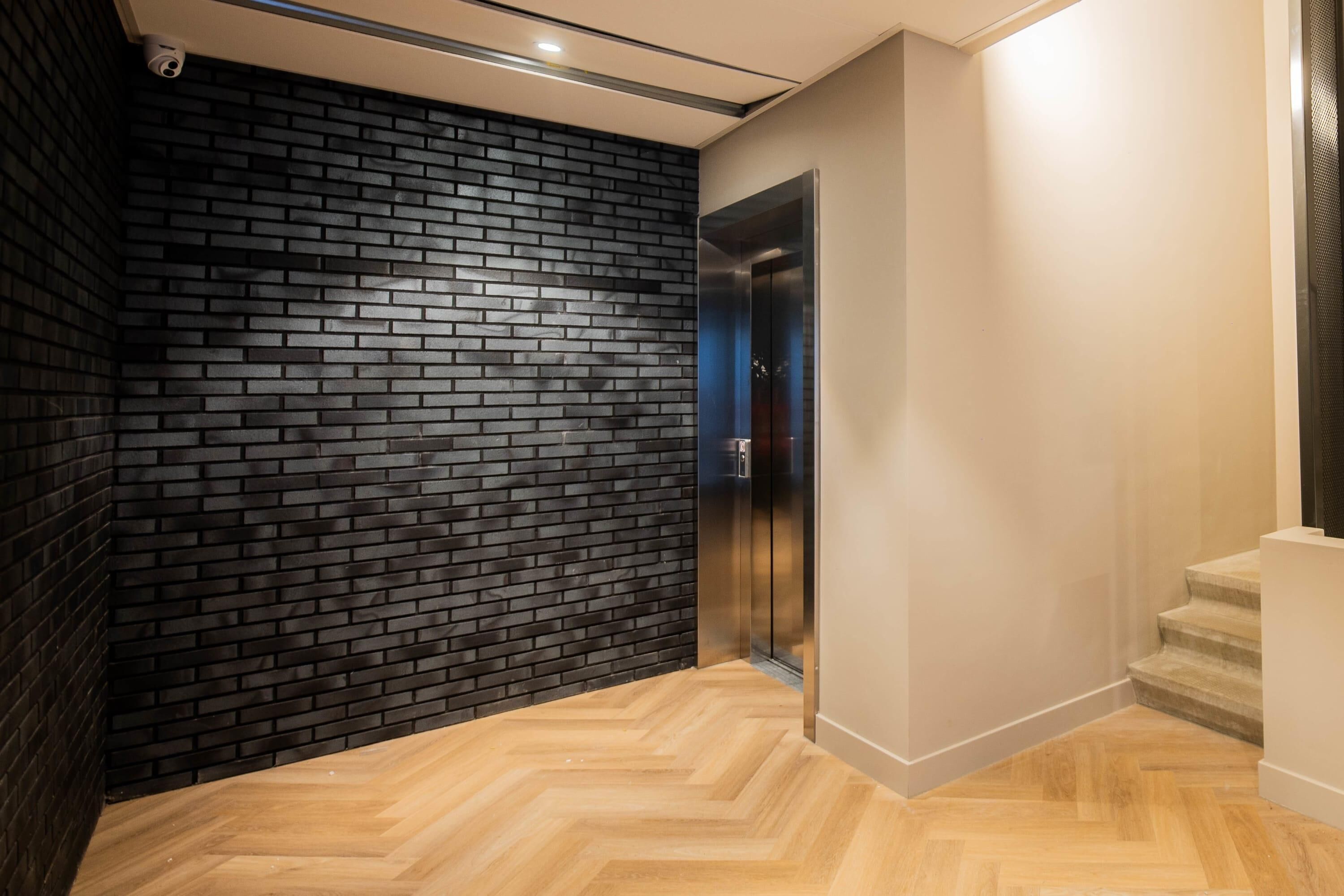 A modern elevator with a metallic finish, adjacent to a black brick wall and a wooden herringbone floor.