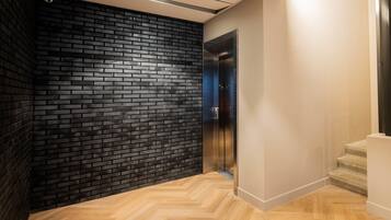 A modern elevator with a metallic finish, adjacent to a black brick wall and a wooden herringbone floor.