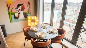 A dining area with a round table set for four, a large window with a view of buildings, and a wall-mounted light fixture spelling out 'B'. - Studio Serviced Apartments With City View (Hilversum)
