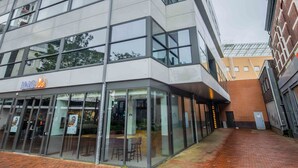 A modern building with a glass facade, a visible entrance, and a brick pathway leading to it. - Studio Serviced Apartments With City View (Hilversum)