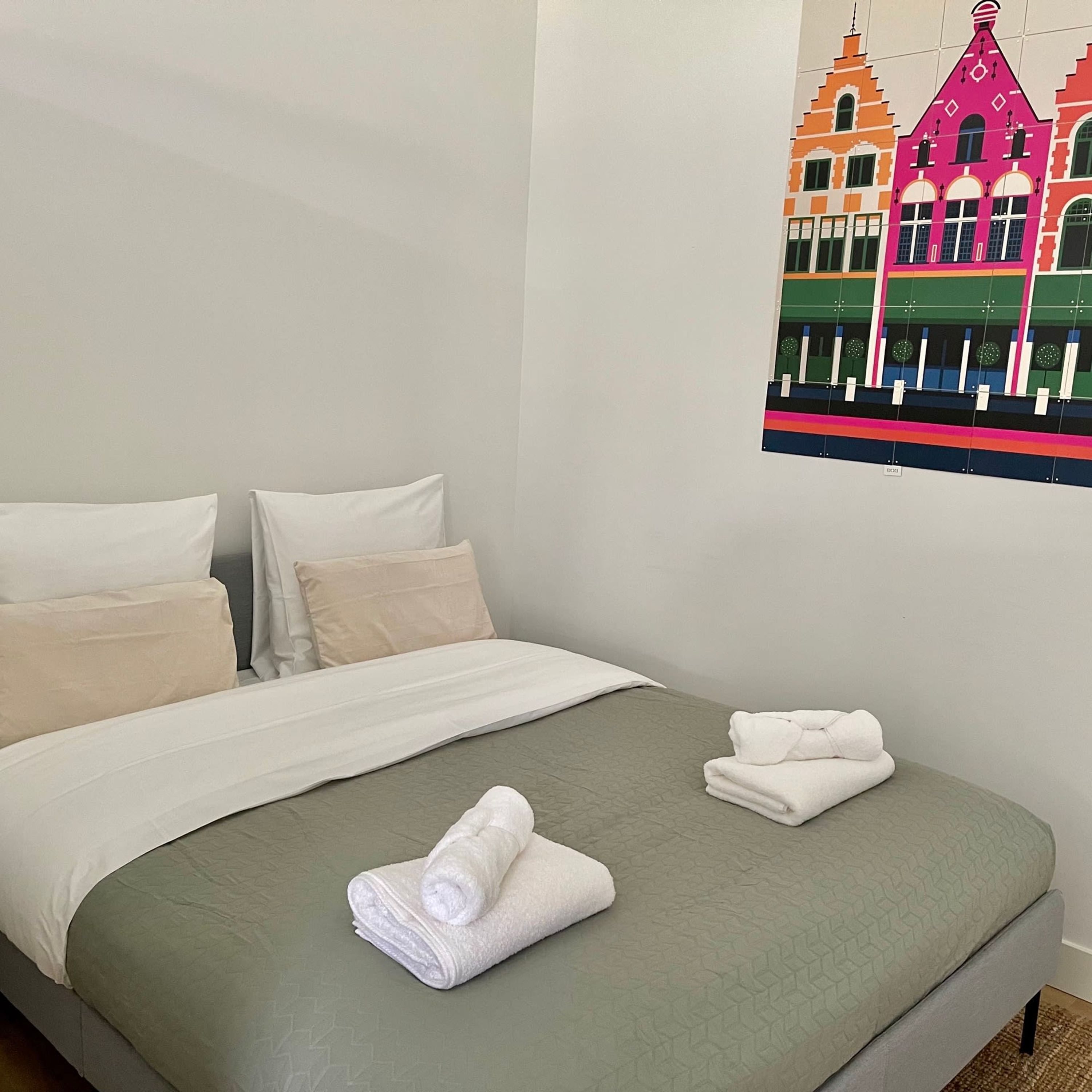 Ein ordentlich bezogenes Bett mit grüner Tagesdecke und weißen Kissen, ein gefaltetes weißes Handtuch auf dem Bett und eine farbenfrohe Illustration eines Gebäudes an der Wand.