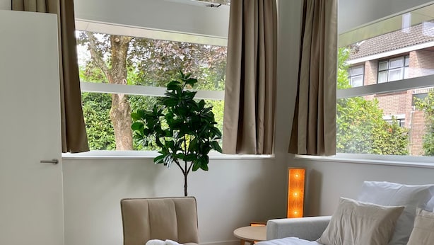 A hotel room with a bed, a chair, a table, a lamp, and a view of trees and buildings through large windows. - Serviced Studio Apartments in Utrecht (De Bilt)