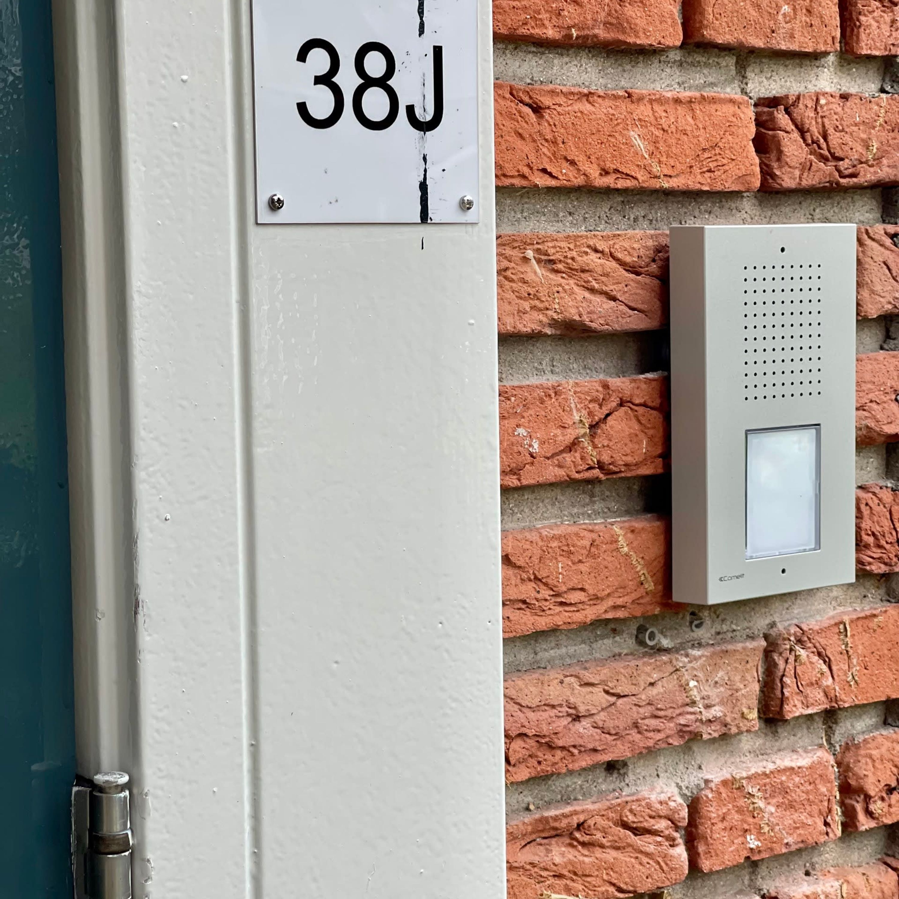 Une porte avec une plaque indiquant « 38J » et un interphone de sécurité fixé sur un mur en briques.