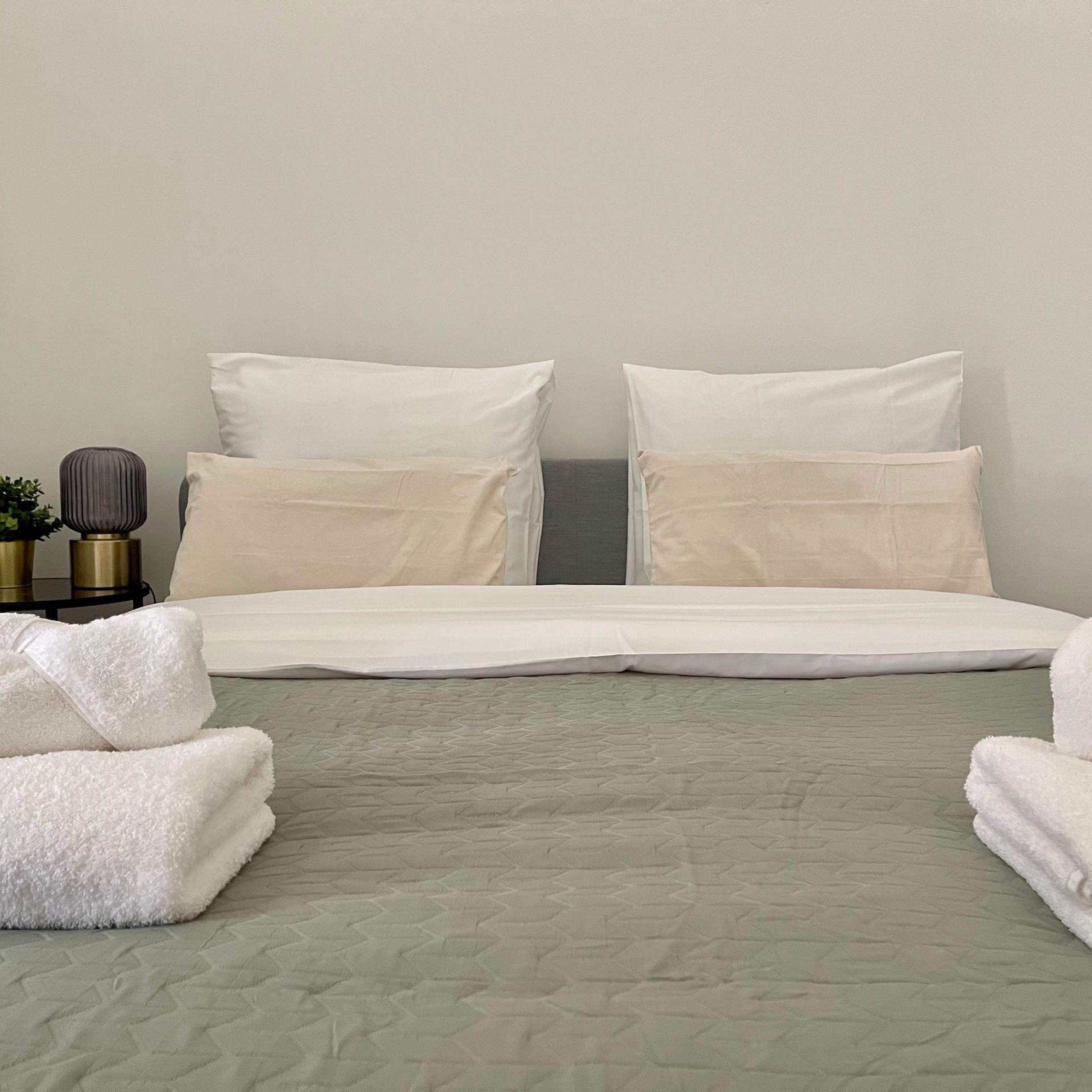Una cama bien hecha con sábanas y almohadas blancas, una mesita de noche con una lámpara y una planta, y dos toallas dobladas en el suelo.
