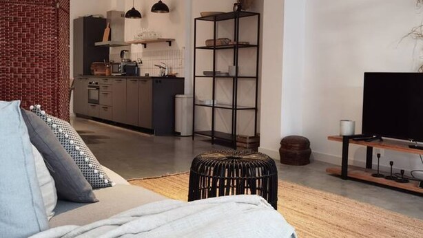 A modern living room with a sofa, ottoman, and a TV stand. There is a kitchen in the background with a dining area.