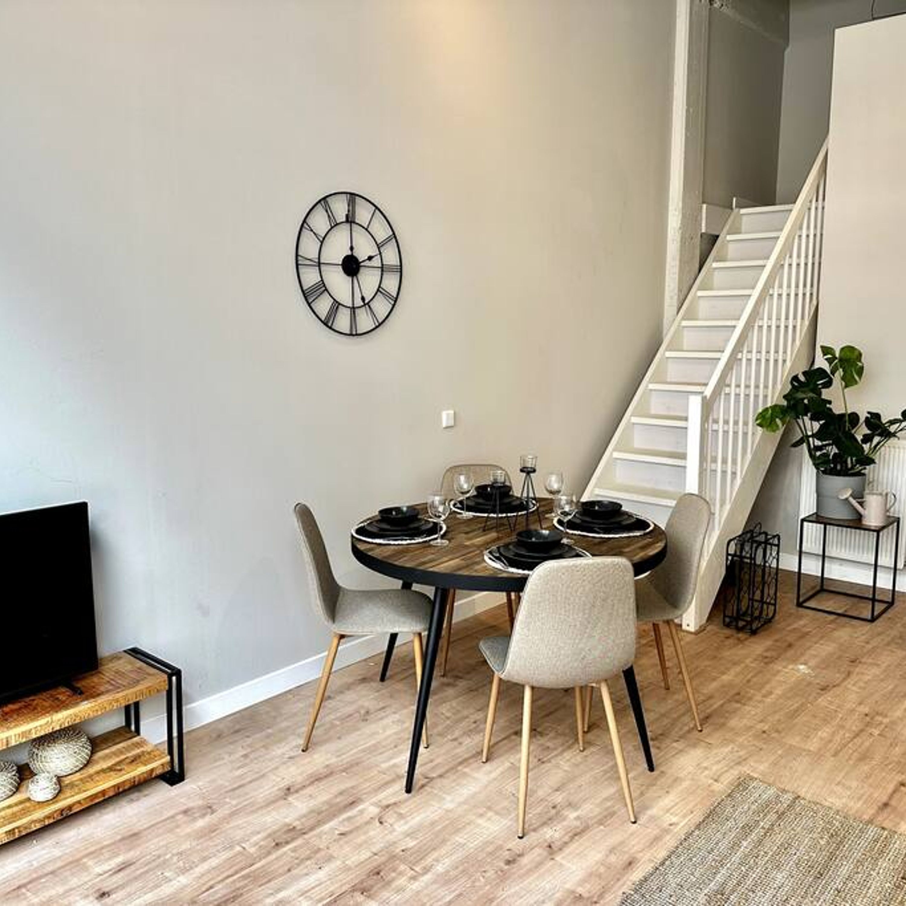 A dining area with a round table, four chairs, a wall clock, and a staircase.