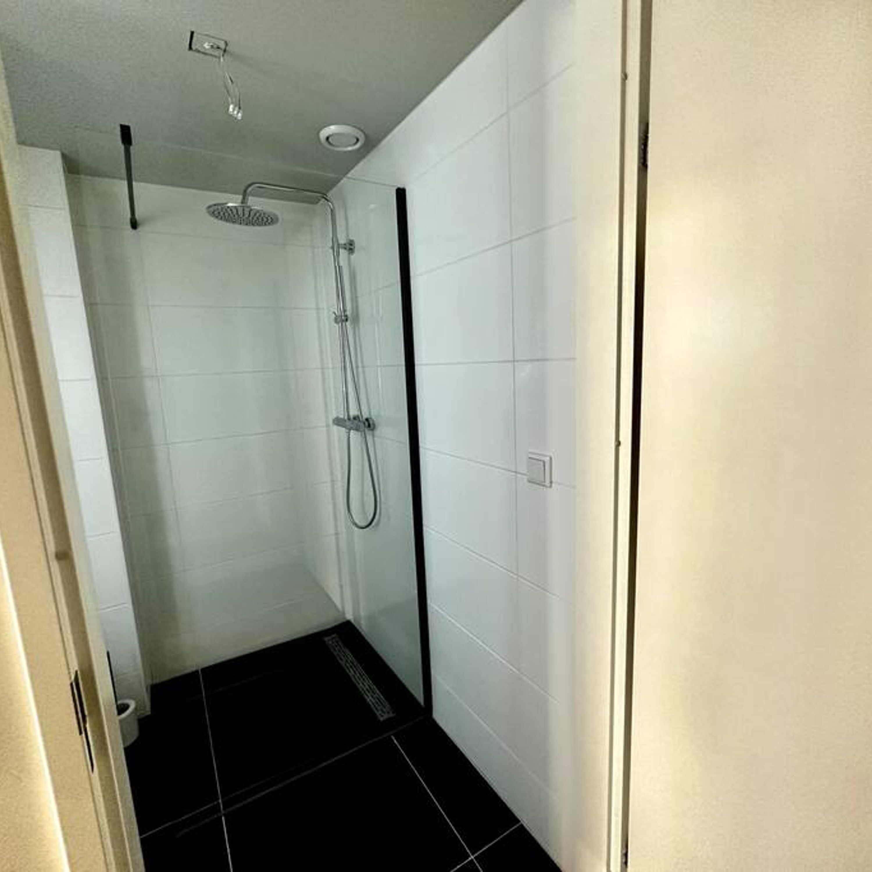 A small, modern bathroom with white tiled walls and a black floor. It includes a shower with a handheld showerhead and a handheld shower attachment.