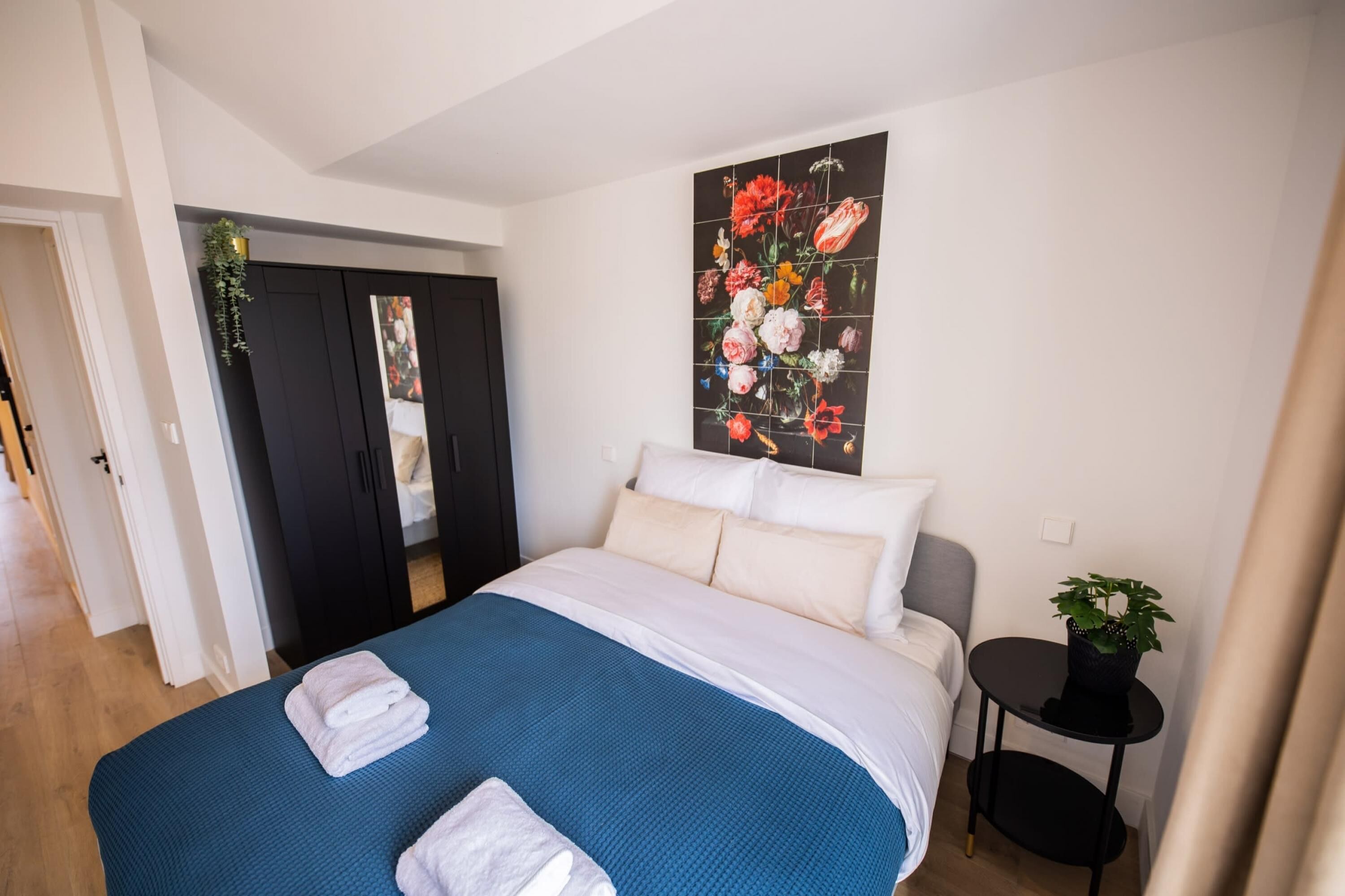 Un dormitorio con cama, almohadas, colcha azul, un cuadro de flores, una mesita con una planta y una puerta que da a otra habitación.