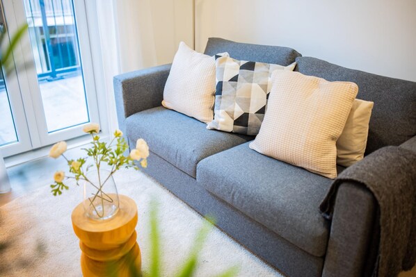 A grey sofa with patterned cushions, a wooden side table with a vase of flowers, and a view of the outdoors through a window.