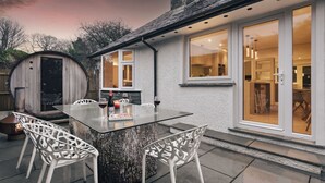 A modern outdoor dining area with a glass-top table, white chairs, and a rustic tree stump base. - Woodlands Family Retreat - Windermere Lakedistrict (Windermere)