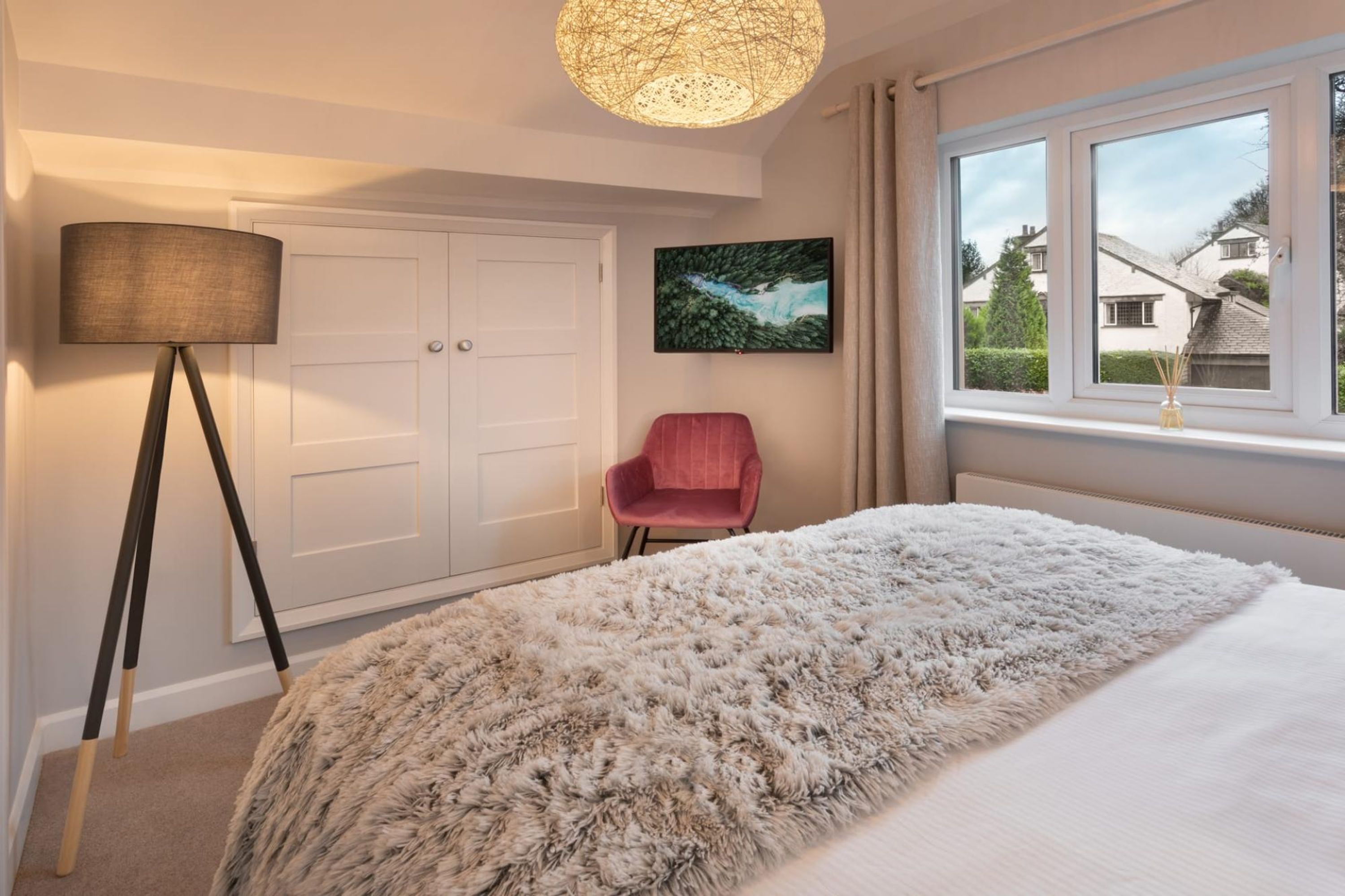 A bedroom with a large bed, a standing lamp, a chair, a painting, and a window with a view of houses.