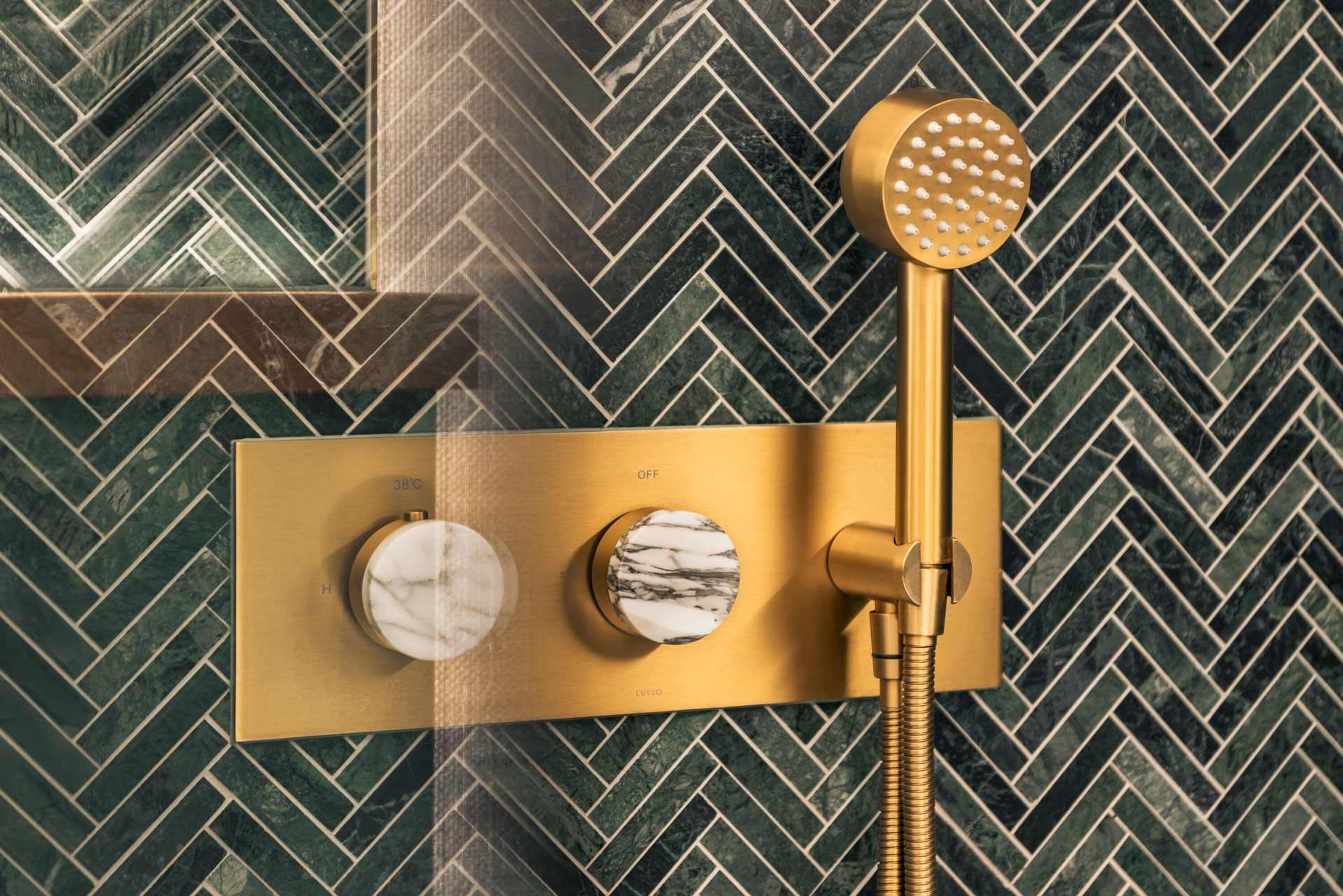 A gold showerhead with a marble-like texture, a gold control panel with a temperature dial, and a herringbone-patterned tiled wall.