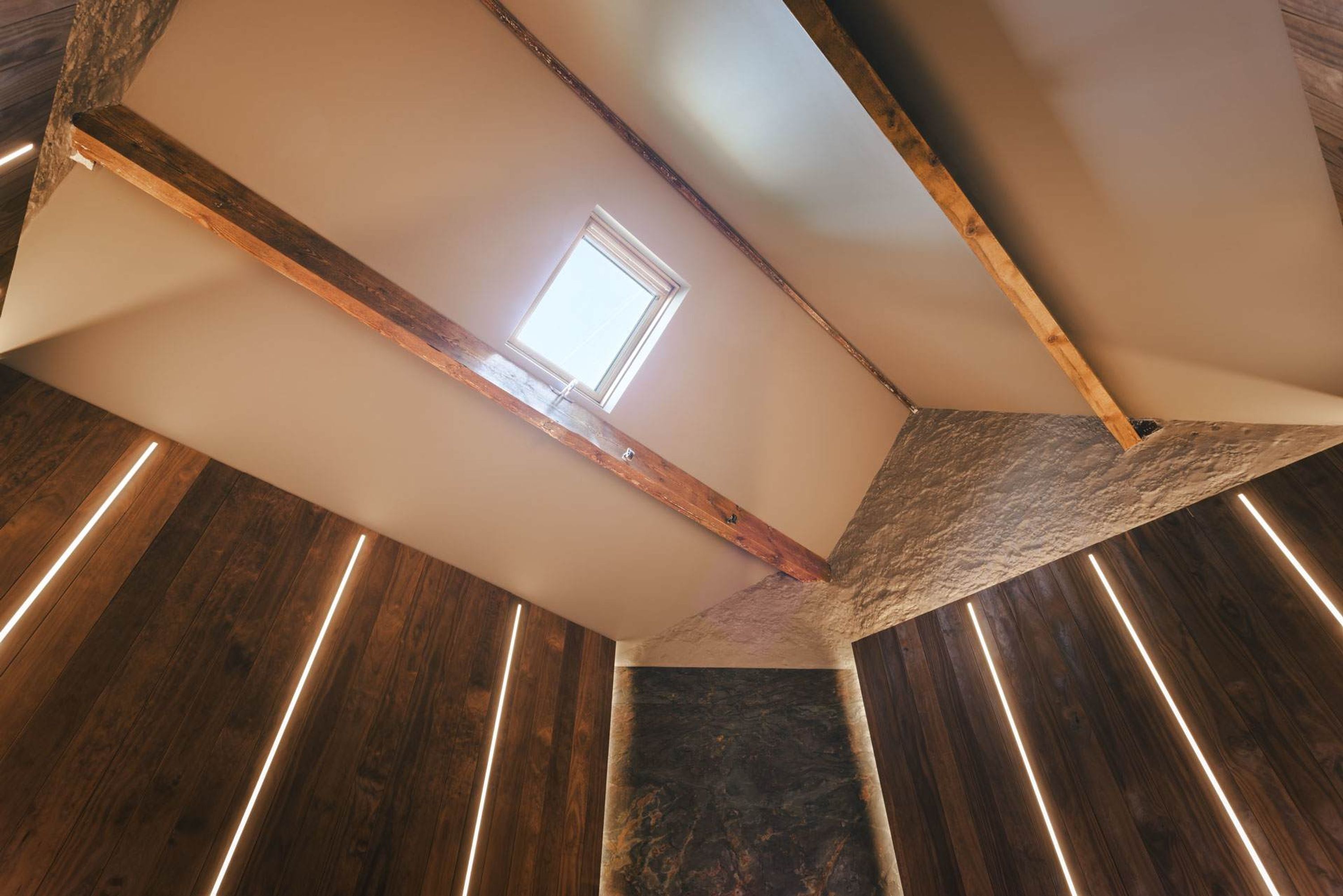 Una habitación con techo inclinado, vigas de madera, tragaluces y suelo de madera.